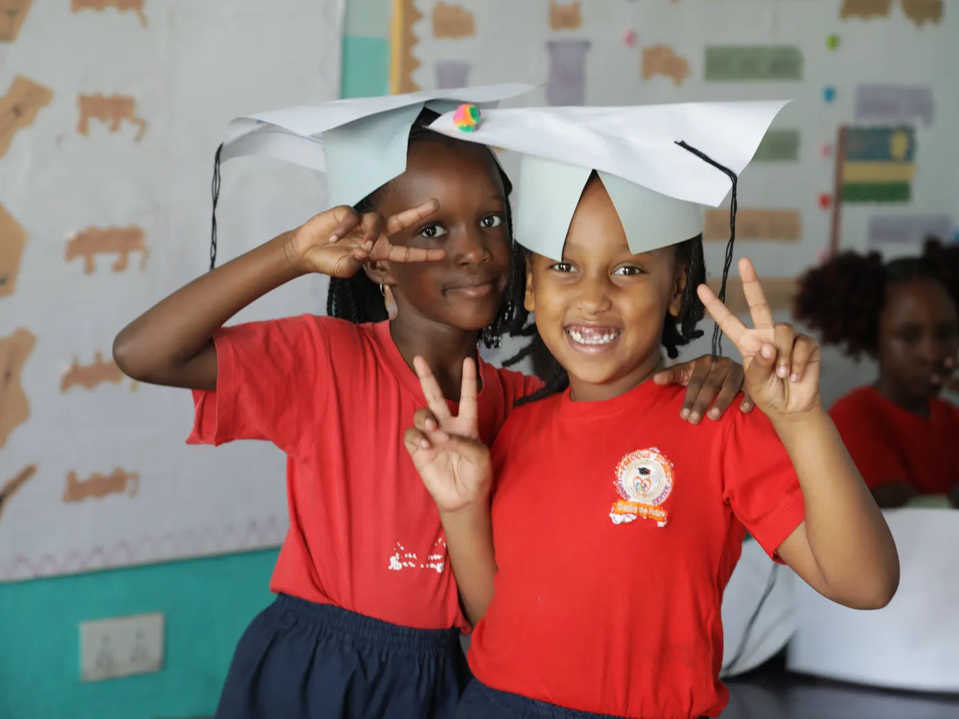 Two pupils laughing in their classroom, wearing paper hats they have made