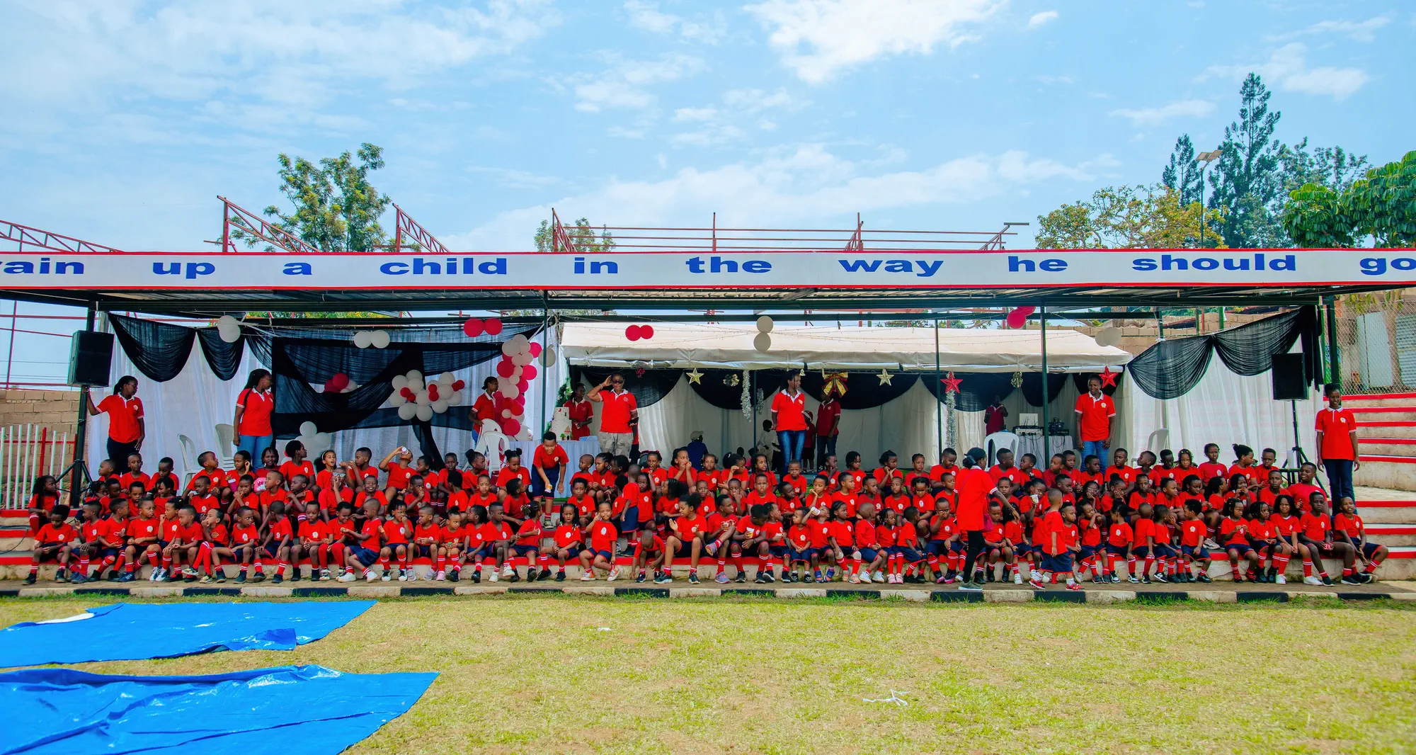 The whole school, pupils and teachers in red, seated together under a banner that reads "Train up a child in the way he should go"