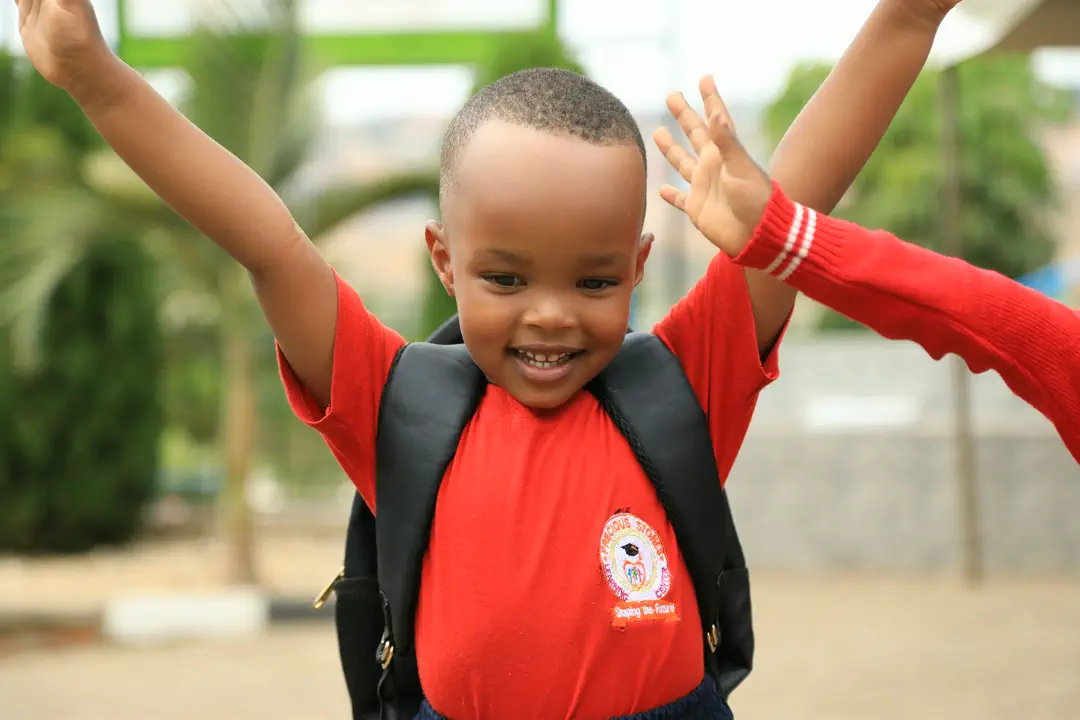 A small boy with a backpack, grinning with his arms flung wide