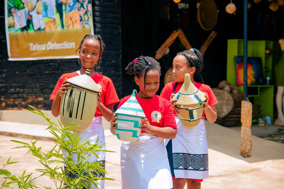 Three girls carrying woven baskets with pointed lids at a cultural village