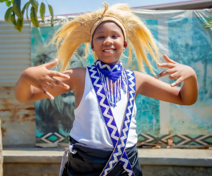 A girl in a straw headdress and beaded sash, smiling in the middle of a traditional dance